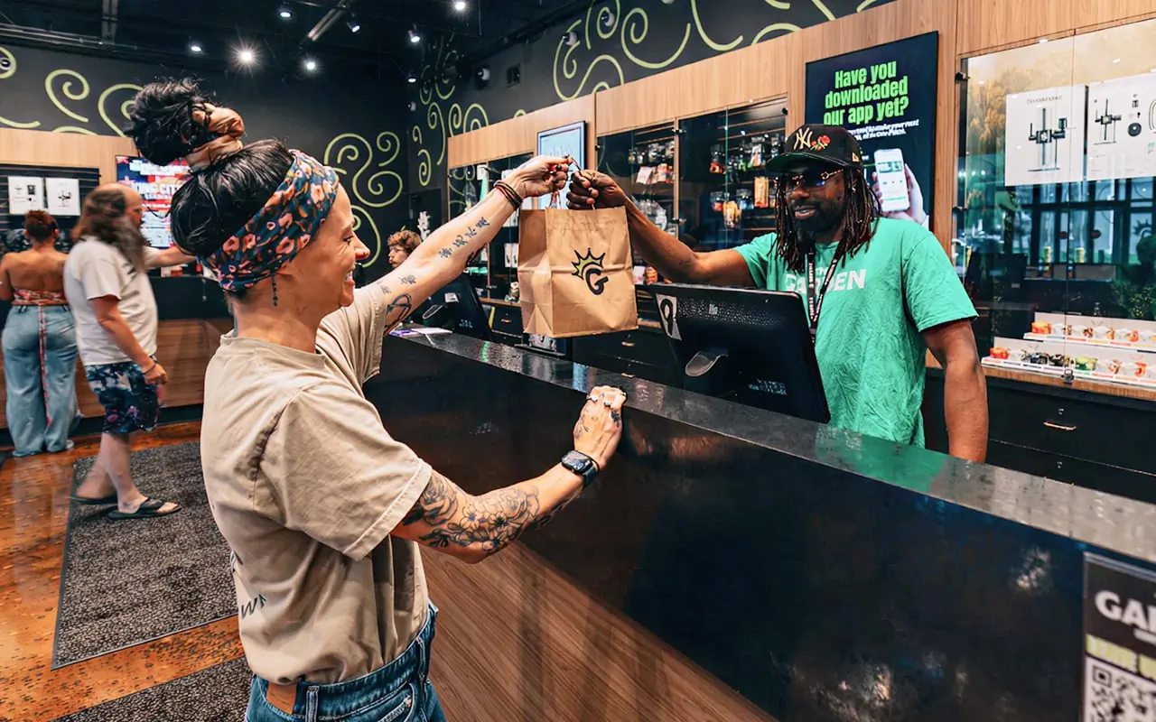A Garden budtender hands a customer a crowned-G bag across the Camp Washington counter